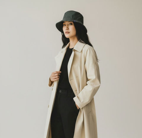 A woman modeling a long beige trench coat over a black outfit, wearing a dark green bucket hat, black pants, and white sneakers, set against a plain light gray studio background.